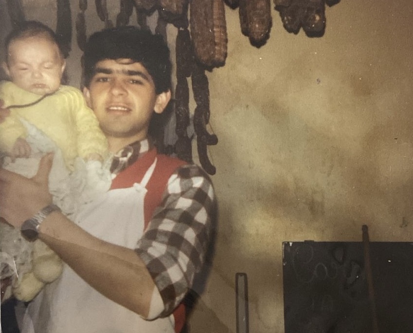 Joven con un bebé en brazos, con embutidos caseros colgados detrás, Arroyo Cabral 1986